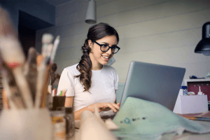 Small business owner woman sitting at desk smiling at laptop with paint brushes and supplies around her.