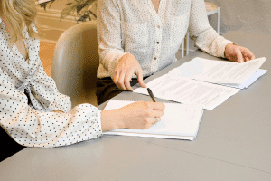 Two women in white blouses signing white sheets of paper on a gray table.