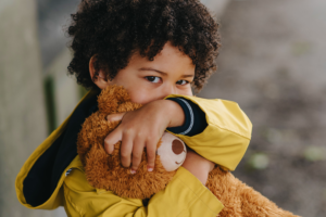A boy with brown curly hair and a yellow rain coat hugging his light brown teddy bear, covering his nose and mouth.