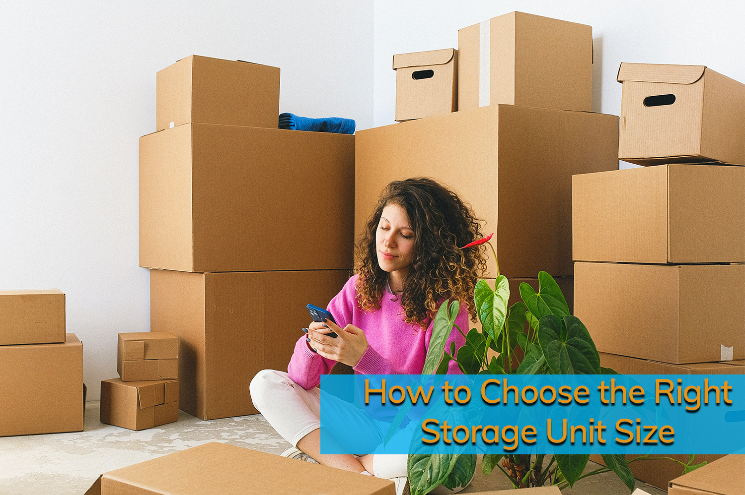 Woman with brown curly hair sitting on floor surrounded by cardboard boxes looking on phone deciding the right storage unit size for her needs.